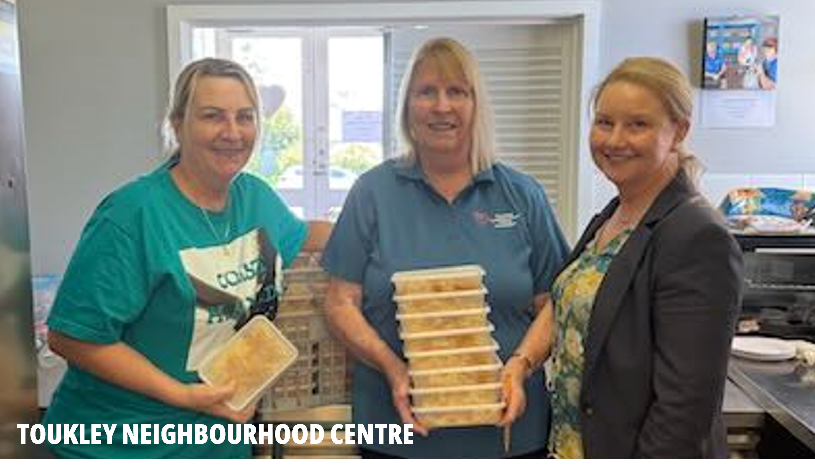 Toukley Neighbourhood Centre picture. Three women stand side by side smiling at the camera. The woman in the middle holds a stack of meals they've prepared in plastic containers.