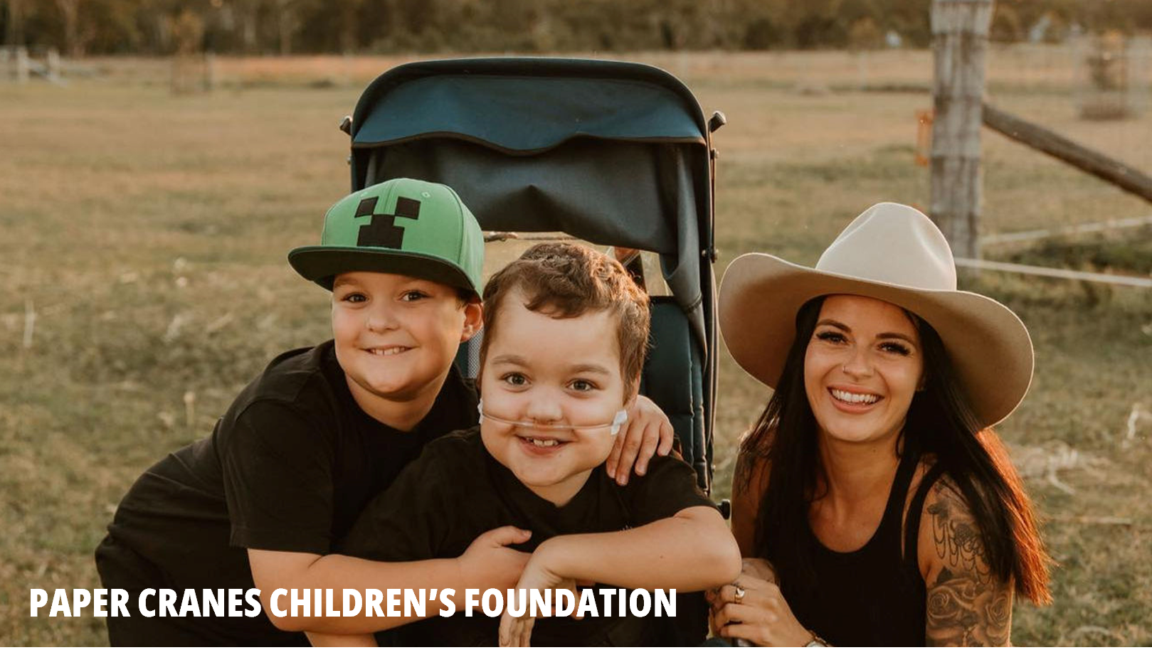 Paper Cranes Children's Foundation picture showing two boys and a young woman in a wide-brimmed hat. The boy on the left is wearing a green Minecraft hat and black t-shirt. The boy in the middle is wearing a black t-shirt and nasal cannula. 