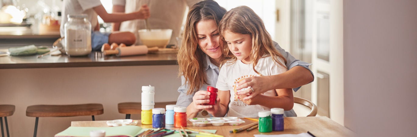 A mother sits with a young daughter at a table covered with paints while in the background a father and son are baking together in the kitchen