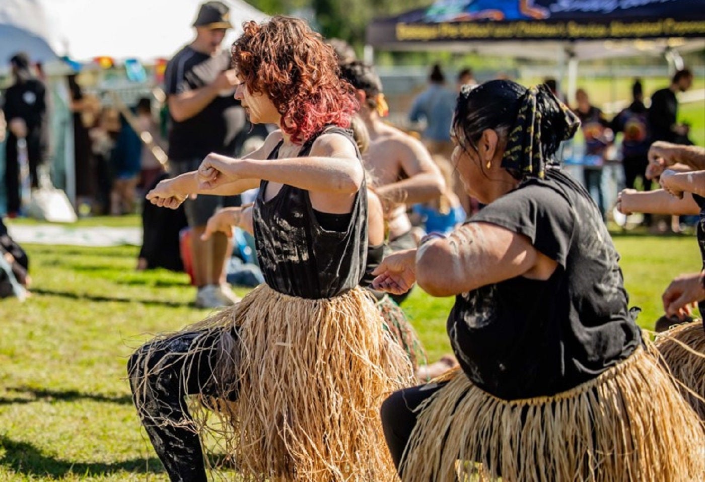 Two First Nations women dancing at the 2024 Saltwater Freshwater Festival in Kempsey, NSW. The woman on the left has red/auburn hair and wears a black vest, black leggings and grass skirt. The woman on the right has dark hair and wears a black t-shirt, black leggings and a grass skirt. Both have white paint on their face and shirts.