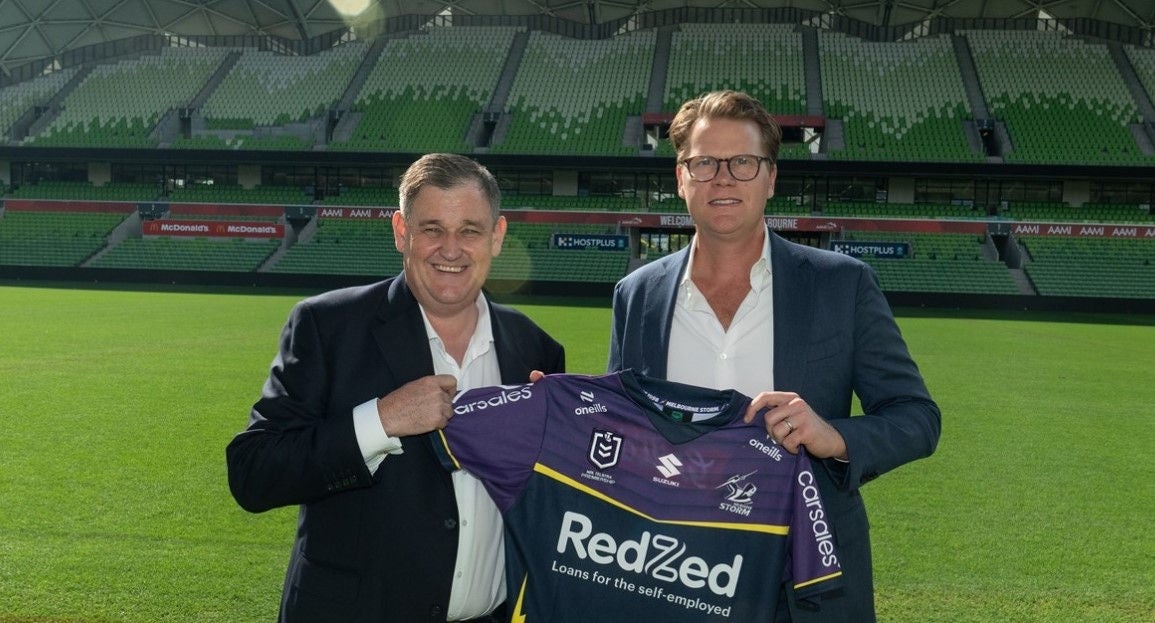 Alinta Energy Managing Director & CEO Jeff Dimery and Melbourne Storm CEO Justin Rodski pose for a photograph at a stadium. They are holding up a Melbourne Storm shirt.