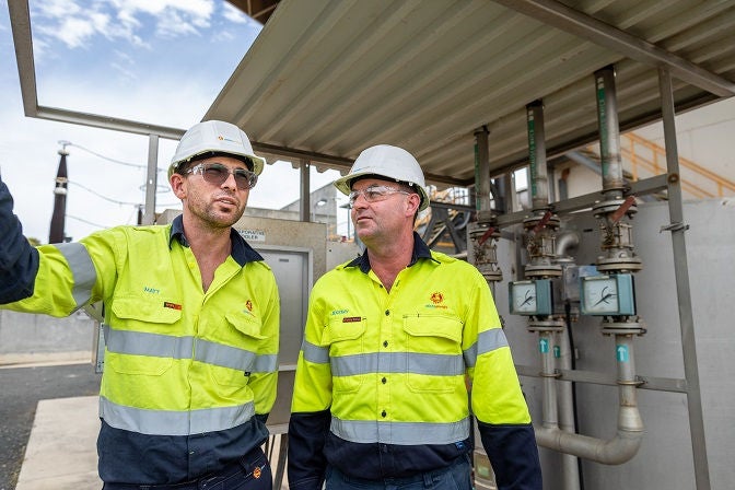 Two Alinta Energy site workers with white hard hats, safety glasses and yellow hi-vis stand next to each other at a power site.