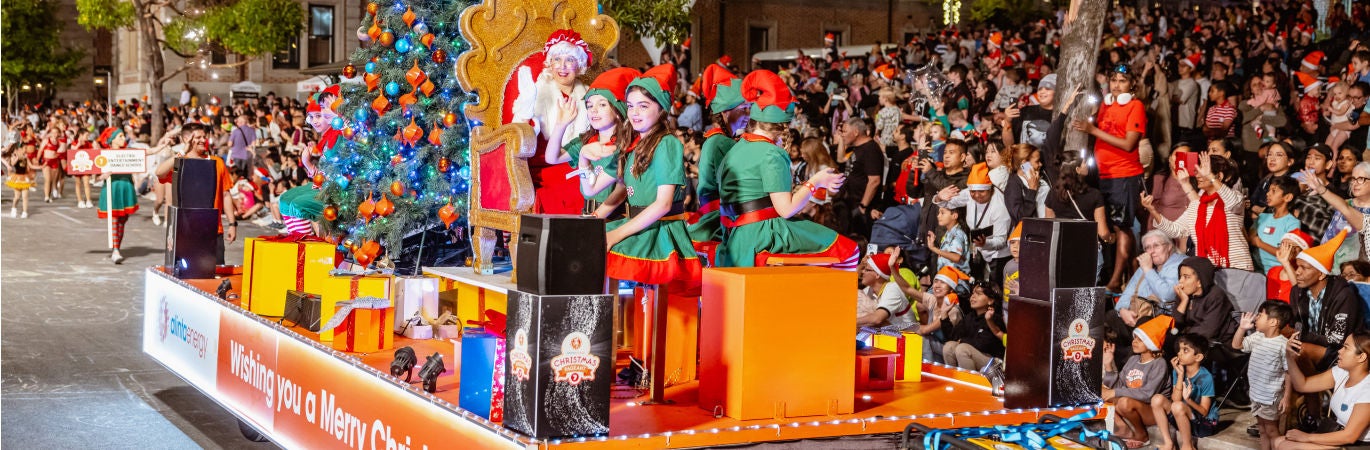 Colourful Christmas float during a pageant, with elves and a Santa, watched by a large crowd