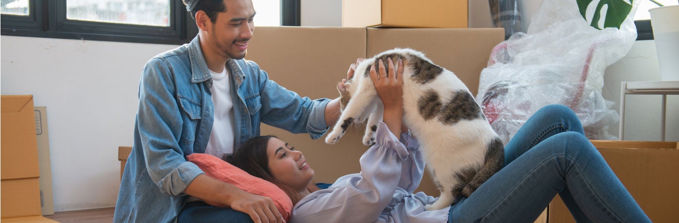 A young couple in their new home, surrounded by moving boxes, lie on the floor and smile and play with their cat