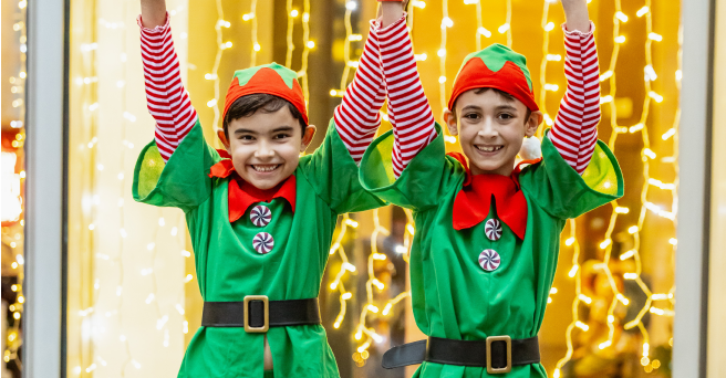 Two smiling children dressed as Christmas elves, holding their hands up in the air