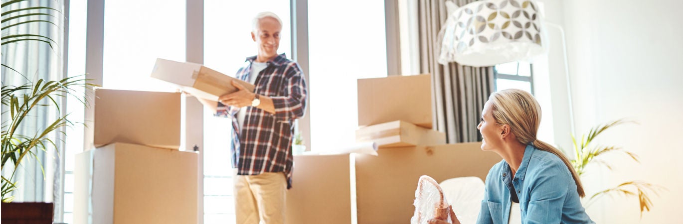 Mature woman sitting on the floor unpacking moving boxes and smiling at mature man who is also unpacking moving boxes in a bright, sunny room