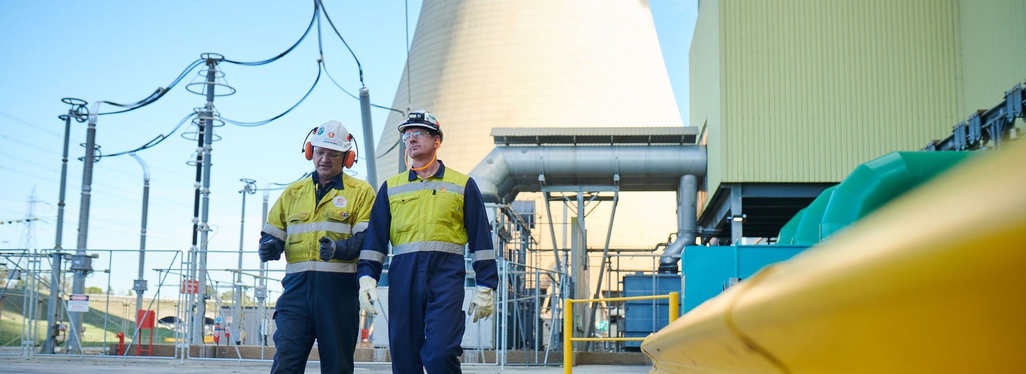 Two workers wearing hi-vis, hard hats and eye protection walk at Loy Yang B Power Station. Behind them is a cooling tower.