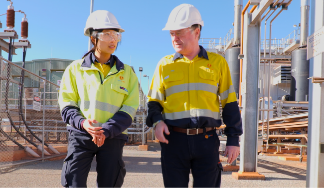 Two Alinta Energy site workers (one male, one female) with white hard hats, safety glasses and yellow hi-vis stand next to each other. They are smiling and looking slightly beyond the camera.