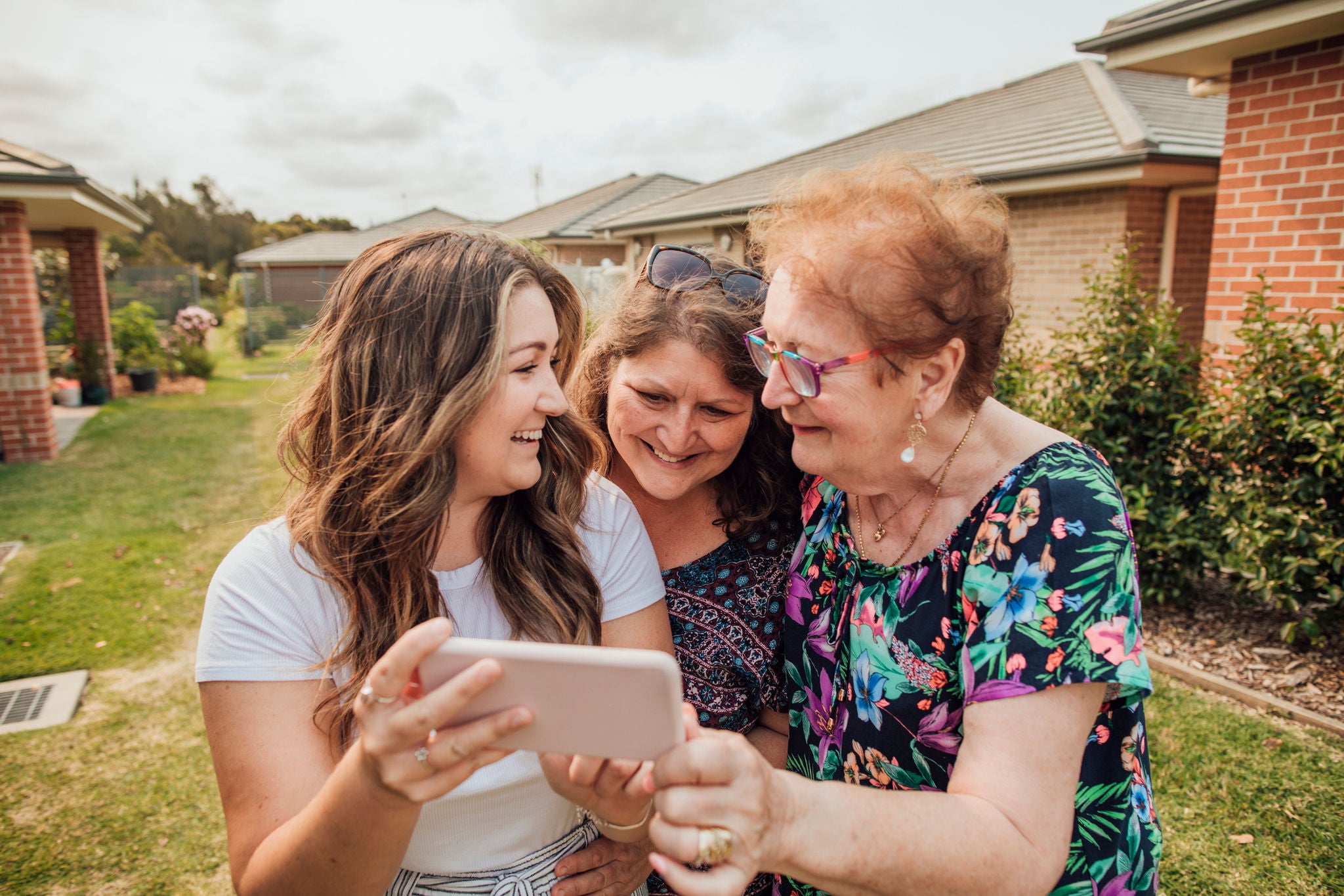 A young woman standing outside some houses shows the picture she has just taken on her mobile phone to two older female family members