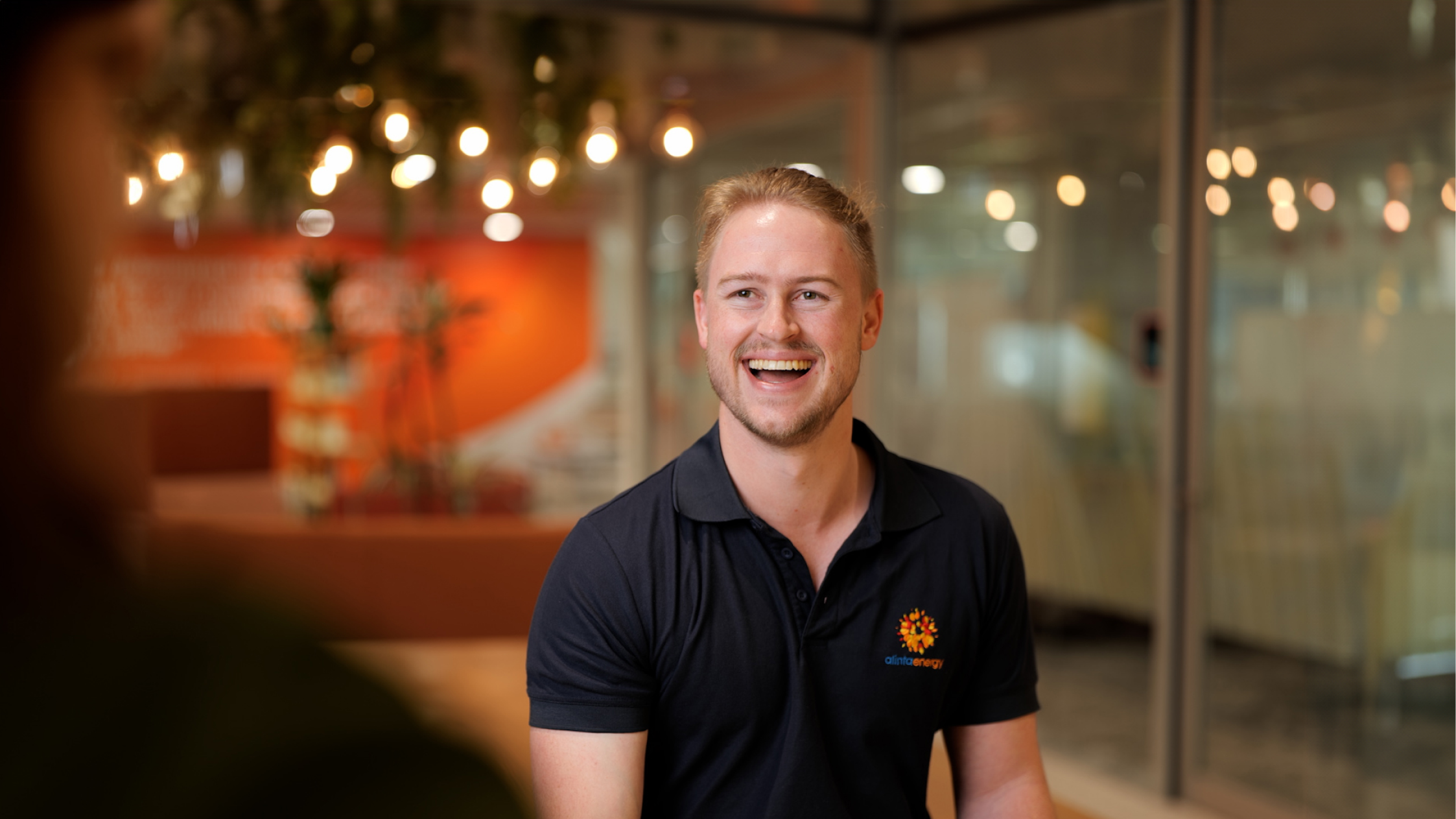 Smiling male Alinta staff member in an open office with glass walls and bokeh hanging lights from the ceiling