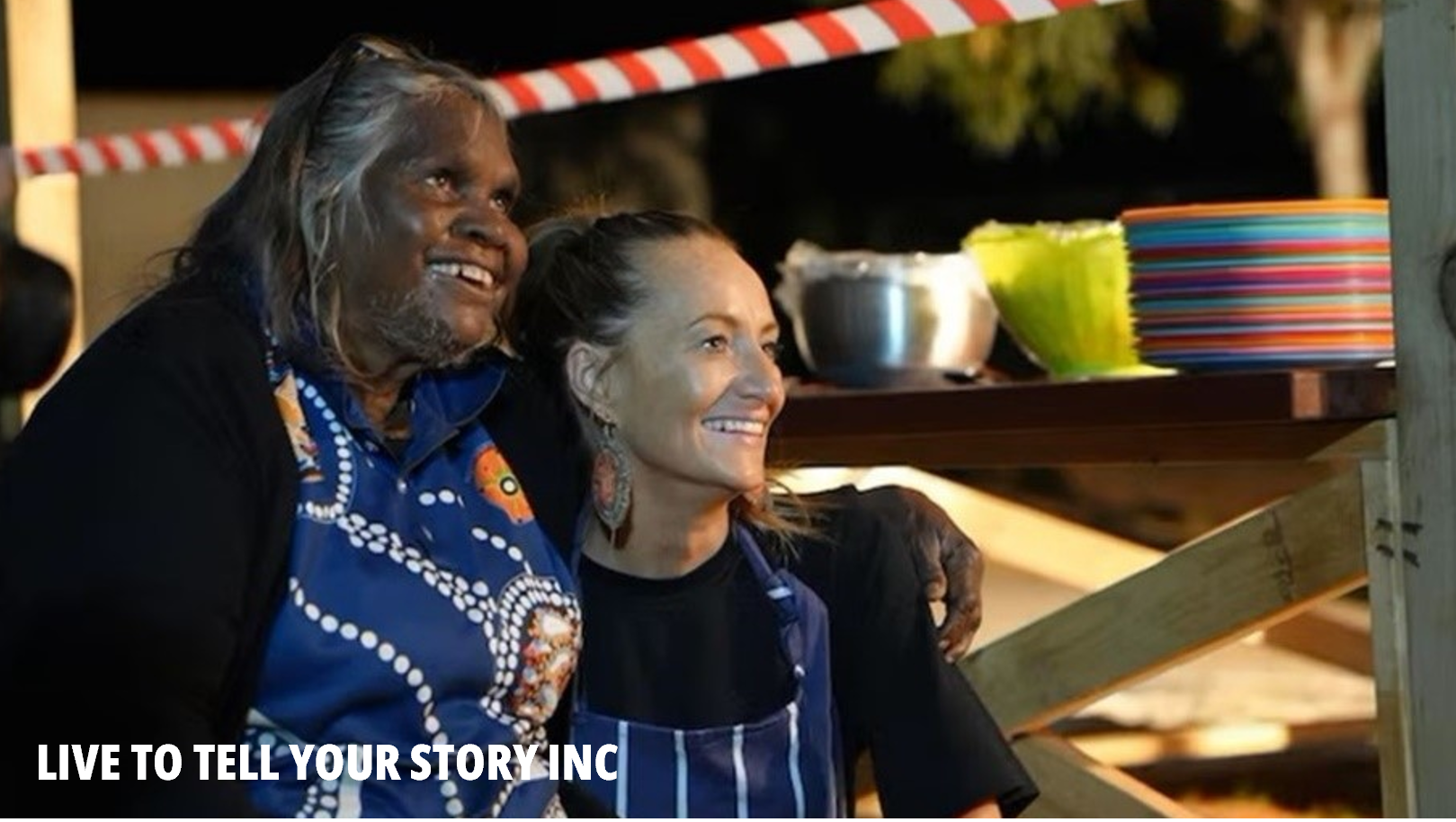 Live To Tell Your Story picture. An older smiling Aboriginal woman with grey-white hair has her arm on the shoulder of a younger woman with pulled-back hair. They are both smiling.