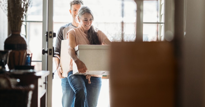 Smiling couple holding a large box, walking in through well-lit open doorway to their new home