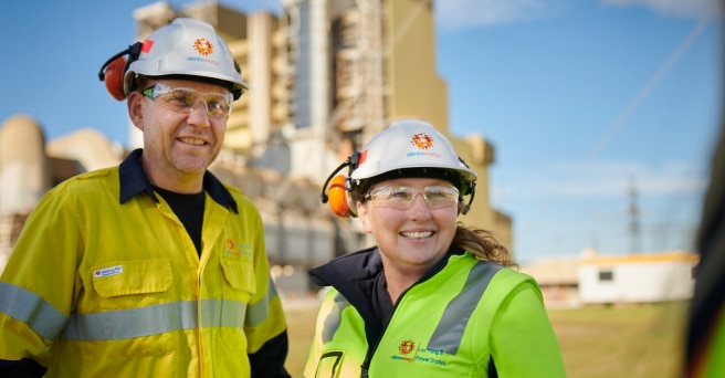 Two smiling workers outside power generating plant, wearing high visibility jackets, hard hats, and ear protectors