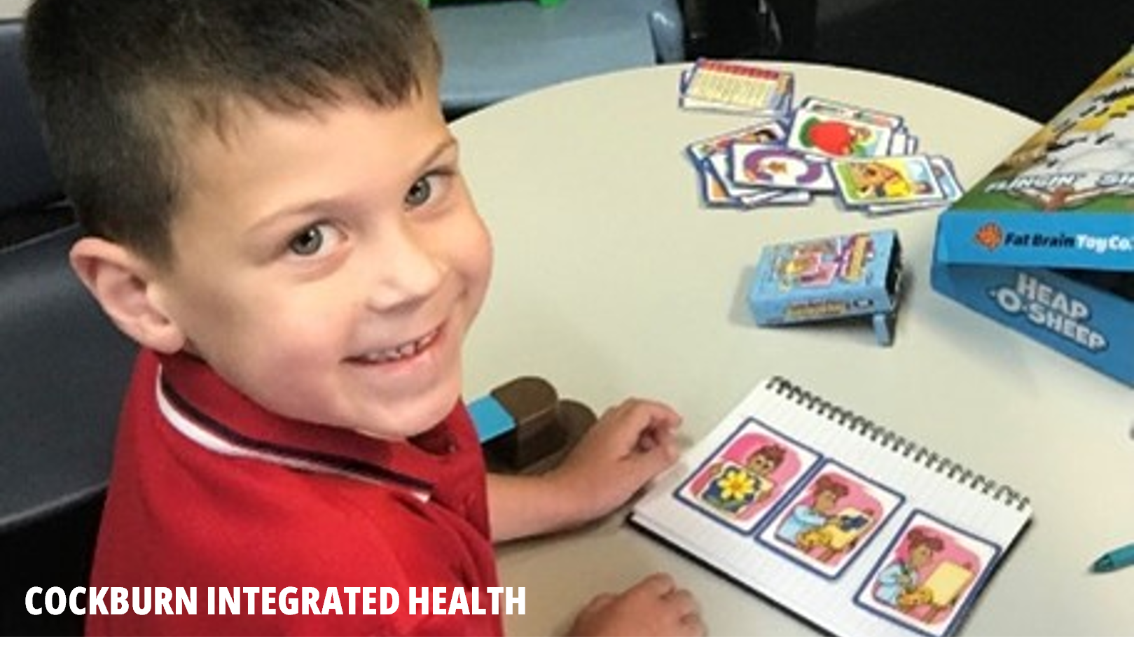 Cockburn Integrated Health photo - an image of a smiling boy in a red top sitting at a table with a card game.