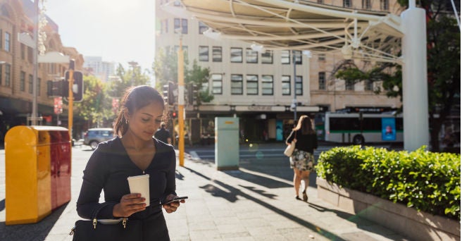 A shot of a businesswoman using her phone and wearing casual business clothing. She is walking in Perth city centre with a coffee in hand on a summer's day.