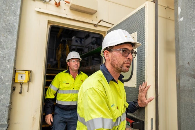 Two Alinta Energy site workers with white hard hats, safety glasses and yellow hi-vis walk through a door on site.