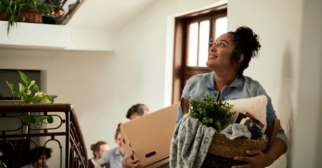 Smiling women carrying a basket of plants and cushions walking upstairs with people carrying moving boxes behind her